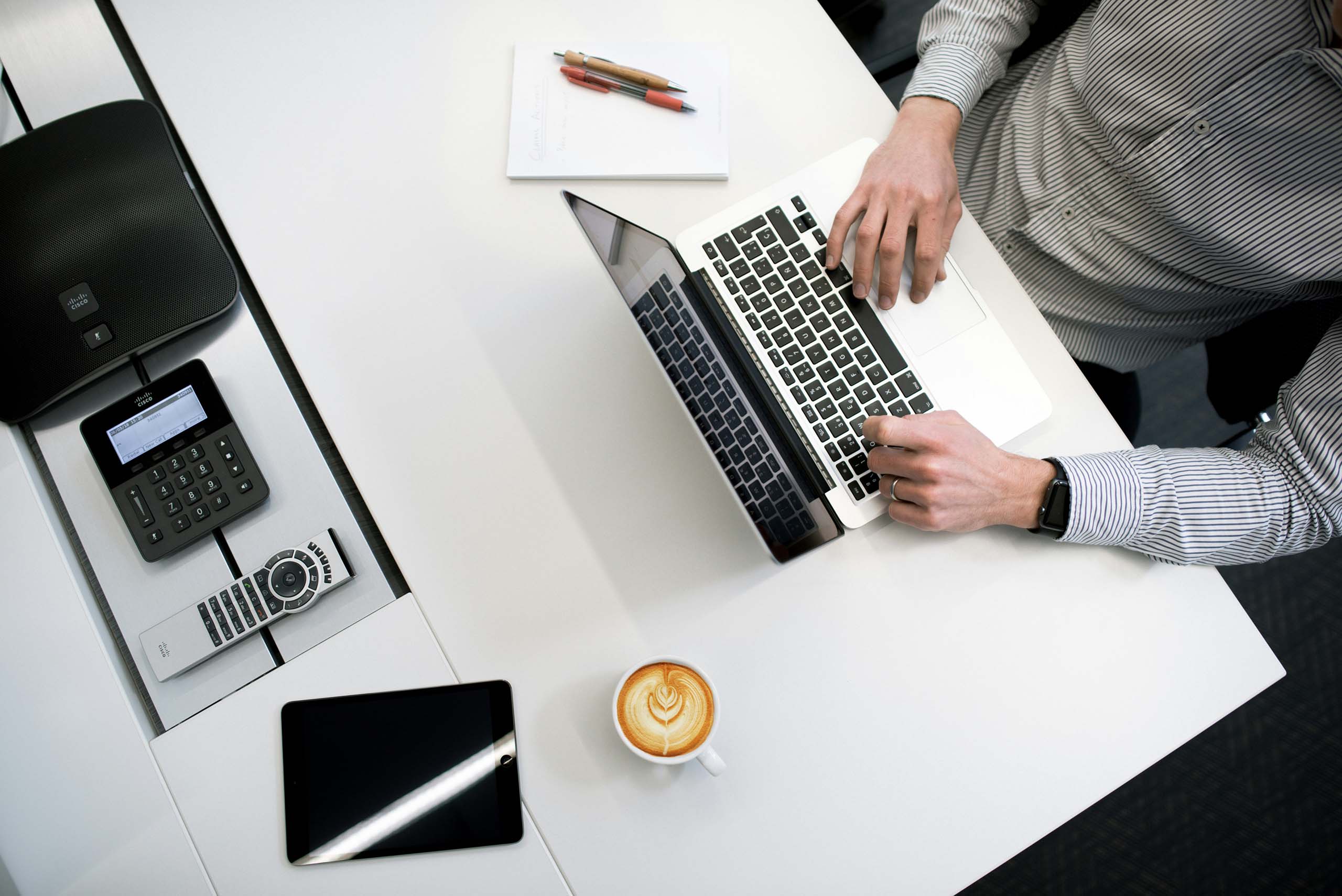 Person am Arbeitstisch mit Laptop, Notizblock, Taschenrechner und Kaffee